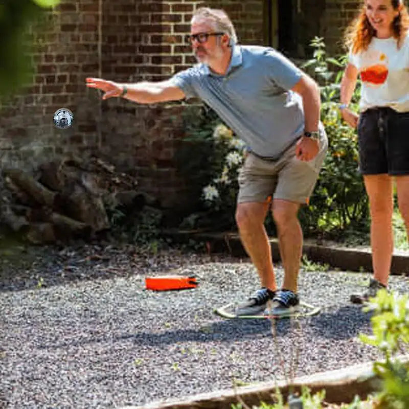 Man playing pétanque with engraved metal balls in a gravel garden, while a woman watches and smiles in the background.